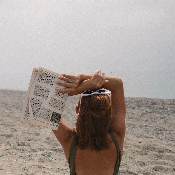 a woman on the beach with white sunglasses on her head looking up at the sun with a crossword in her hand.