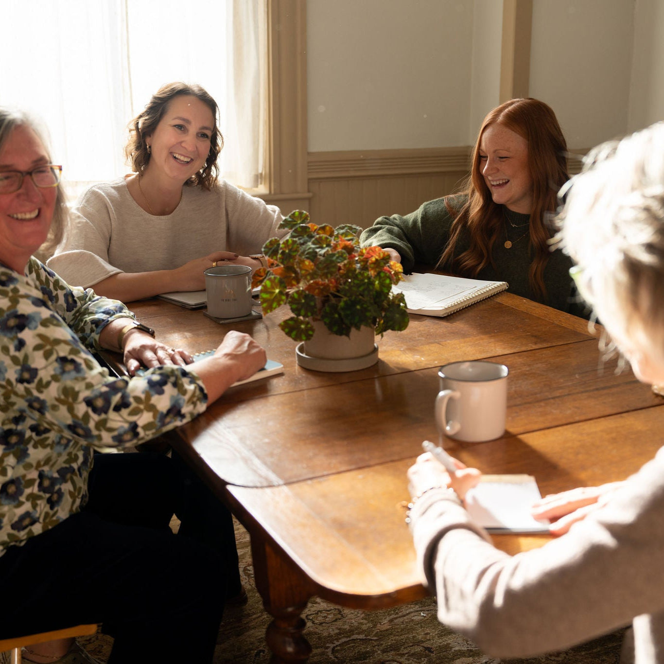 A team of women around a wooden table with flowers on it having a morning meeting with coffee and notebooks out.